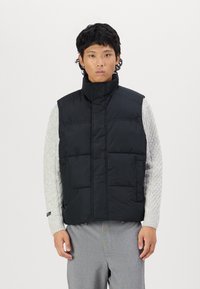 Vest puf negru cu guler înalt, design matlasat, buzunare frontale, purtat peste un pulover texturat gri deschis, asociat cu pantaloni gri.