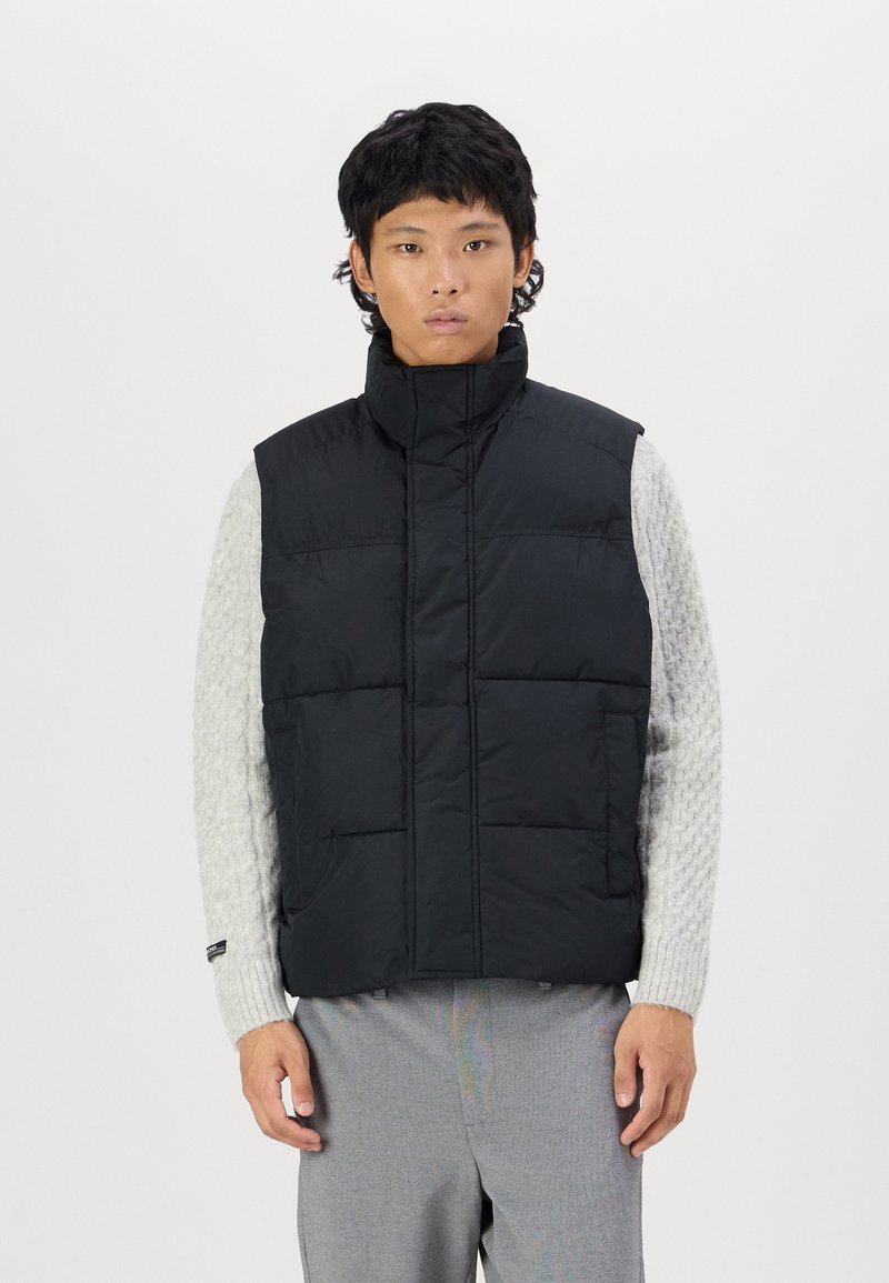 Vest puf negru cu guler înalt, design matlasat, buzunare frontale, purtat peste un pulover texturat gri deschis, asociat cu pantaloni gri.