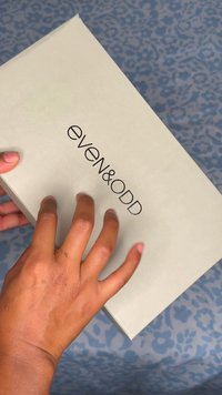 Dłoń trzymająca szary pojemnik z napisem "EVEN & ODD" na tle niebieskiego wzorzystego materiału.