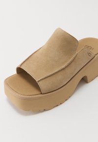 Beige suede platform slipper sandaal met een brede bovenste band en een gestructureerde zool. Het ontwerp heeft zichtbare stiksels langs de randen.
