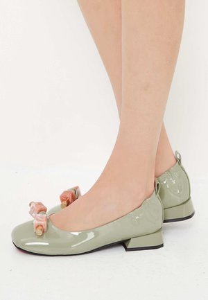 Chaussures vert clair brillantes à talon bas, avec des ornements décoratifs en bloc couleur pêche, portées sur des pieds croisés sur fond blanc.