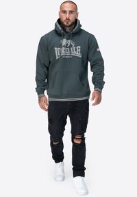 Sudadera verde oscura con el logo de Lonsdale London, bolsillo canguro y capucha con cordón; combinada con jeans negros desgastados y zapatillas blancas.