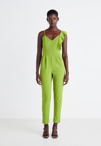 Žalias šokinėjimo kostiumas su V formos iškirpte, viena raukinėta petneša, prigludusia juosmeniu ir siaurinančiomis kojomis. Jame naudojama lygi medžiaga ir klostės priekyje.