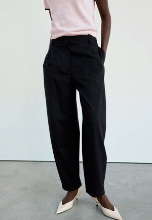Persona que lleva pantalones negros a medida de cintura alta con bolsillos, top rosa claro de manga corta y tacones bajos puntiagudos blancos.