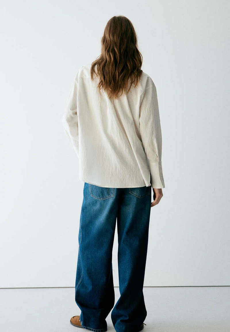Personne aux cheveux longs portant une chemise blanche ample et un jean bleu large, debout face à un mur blanc uni.