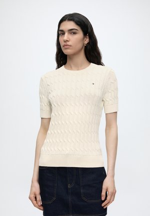 Kvinde med langt mørkt hår iført en cremefarvet kortærmet kabelstrikket sweater og mørk denimskørt, stående mod en ensfarvet hvid baggrund.