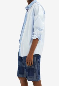 Camisa de botón a rayas azul claro con mangas remangadas, combinada con pantalones cortos navy estampados que presentan diseños geométricos en azul más claro.