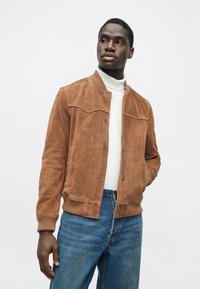 Moški nosi rjavo antilopovo jakno čez belo turtleneck majico in modre kavbojke, nekoliko obrnjeno na stran proti enotni beli ozadju.