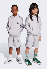 Set gri format din hanorac și pantaloni scurți cu dungi laterale, logo brodat și buzunar frontal. Încălțăminte albă și verde. Șosete cu dungi la ambele picioare.