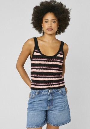 Femme avec une coiffure afro portant un débardeur en maille rayé noir et rose et un short en jean bleu ample, debout, les mains dans les poches.