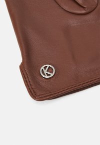Braunes Lederaccessoire mit einem ausgefallenen silbernen kreisförmigen Beschlag mit einem "K"-Logo, das eine glatte Textur und präzise Nähte aufweist.