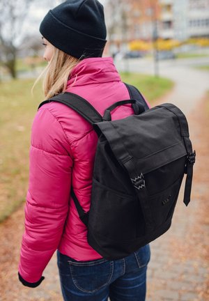Black backpack with a flap and buckle closure, made of durable material. The user wears a bright pink puffer jacket and a black beanie.