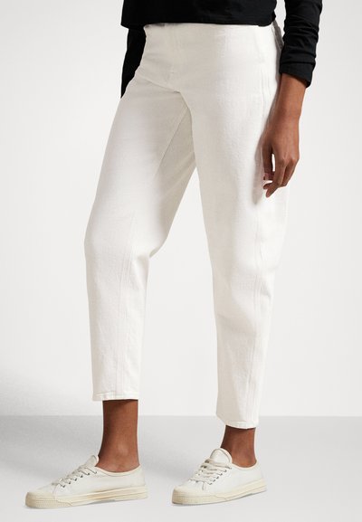 Person trägt weiße, enganliegende, knöchellange Hosen und hellbeige Schnürsneaker und steht vor einem einfarbigen Hintergrund.