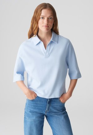 Light blue polo shirt with a collar, short sleeves, and a relaxed fit, paired with light blue jeans. Features a smooth texture.