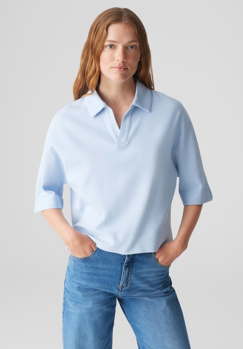 Helle blaue Polohemd mit Kragen, kurzen Ärmeln und lockerer Passform, kombiniert mit hellblauen Jeans. Hat eine glatte Textur.
