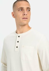 Crème-kleurig langemouwen shirt met een ronde hals, drie zwarte knopen en een borstzak aan de linkerkant. Zachte, gladde stof.