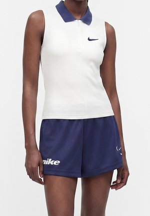 Vrouw draagt een witte mouwloze polo met een marineblauwe kraag en marineblauwe Nike-short met logo's op de linker dij en rechter zoom.