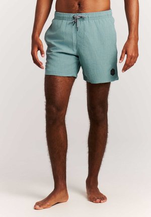 Homme portant un short de bain bleu clair avec un logo noir sur la jambe gauche, debout pieds nus devant un fond uni.