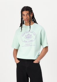 Young man with long braided hair wearing a light green oversized New Era Cap Co New York t-shirt and black pants, hands in pockets.