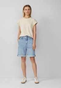 Beige t-shirt met korte mouwen, gecombineerd met lichtblauwe denim shorts; shorts hebben opgerolde cuffs. Witte sneakers met tan accenten maken de outfit compleet.