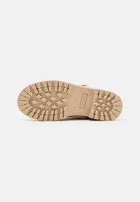 Semelle de botte en caoutchouc beige avec un motif de bande de roulement texturé, comportant une étiquette de marque, des grips ronds et en forme de losange pour une traction optimale.