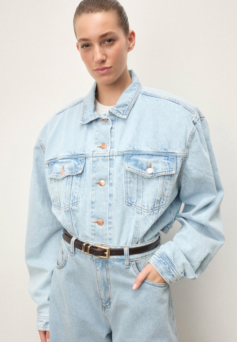 Kobieta z włosami związanymi do tyłu, ubrana w jasnoniebieską, luźną kurtkę dżinsową i pasujące jeansy, czarny pasek, ręka w kieszeni, neutralne tło.