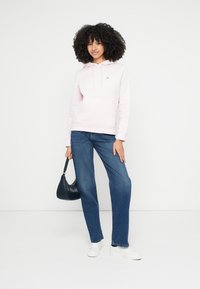 Jasnoróżowa bluza z kapturem z przednią kieszenią, zestawiona z niebieskimi dżinsami o prostych nogawkach oraz białymi trampkami. Trzyma małą czarną torebkę.