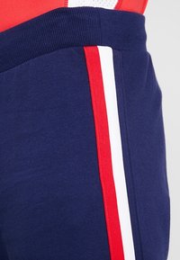 Granatowe spodnie sportowe z grubym elastycznym pasem. Posiadają pionowy detal w czerwono-białe paski po boku dla kontrastu. Gładka tekstura.