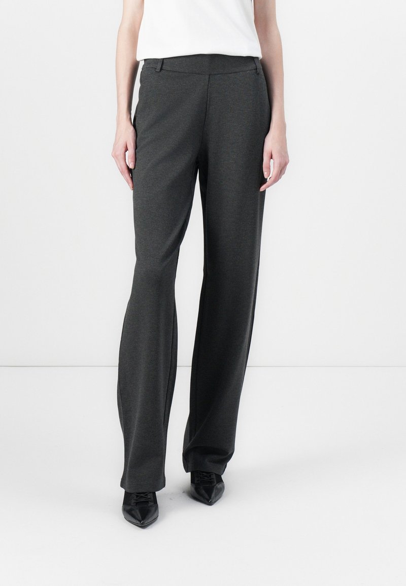 Personne portant un pantalon large gris foncé associé à des chaussures noires à bouts pointus et un haut blanc, se tenant devant un fond blanc.