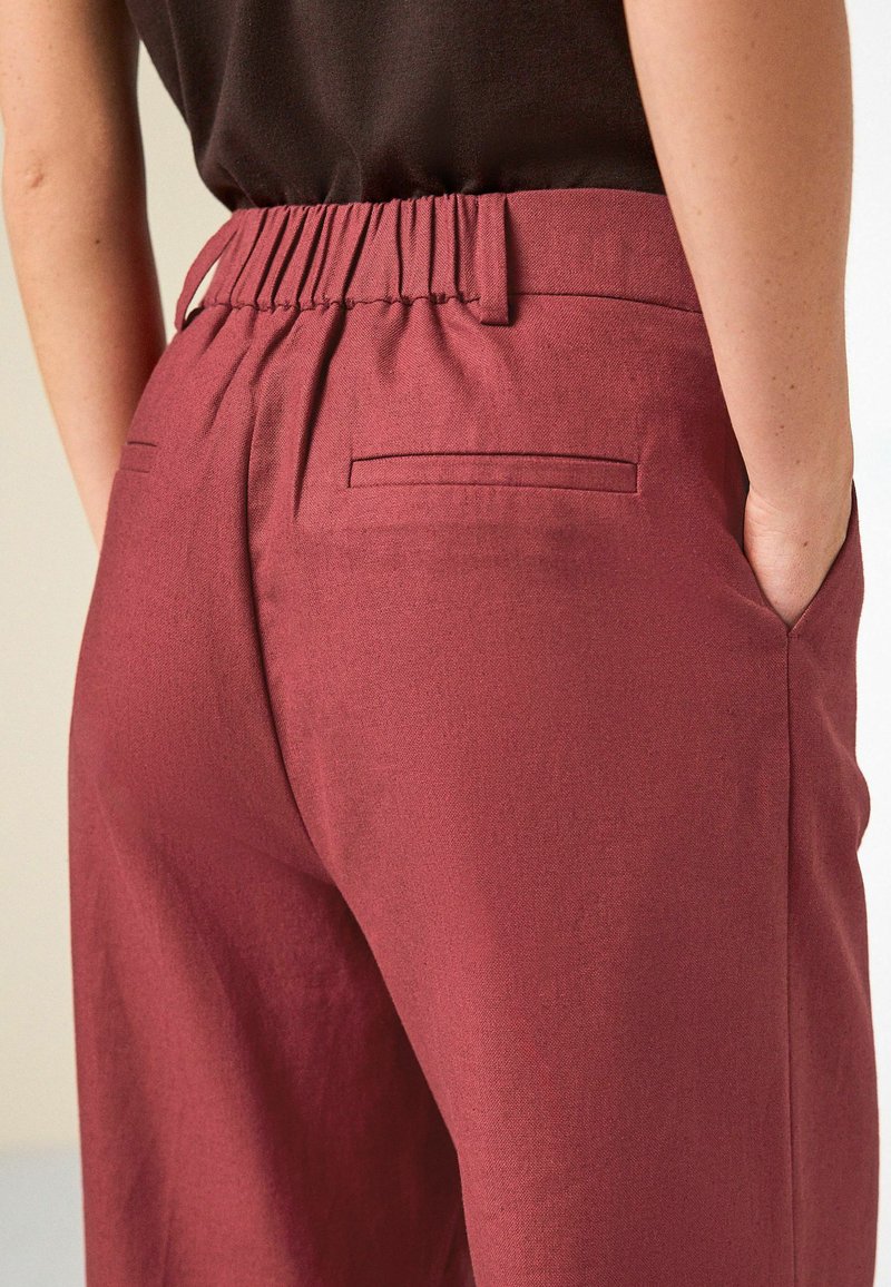 Persona que lleva pantalones de talle alto de color rojo oscuro con cintura elástica y bolsillos de ojal traseros, mano apoyada en el bolsillo lateral, top negro visible.