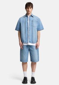 Chemise en denim bleu clair à manches courtes, fermeture par boutons et poche poitrine, associée à un short coordonné jusqu'aux genoux et des chaussures noires.