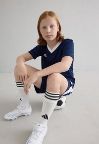 Tumesinine jalgpallipaita valge V-kaelusega, kombineeritud tumesiniste lühikeste pükstega. Valged sokid mustade triipudega. Valged jalgpalliketsid. Istub jalgpallil.