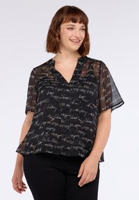 Femeie zâmbind, îmbrăcată cu o bluză neagră transparentă, cu un print de text deschis la culoare și mâneci scurte, combinată cu pantaloni negri.