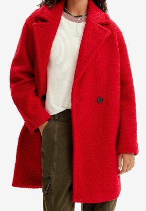 Personne portant un manteau en laine rouge vif sur un pull blanc côtelé et un pantalon cargo vert olive, avec un collier cœur en argent.