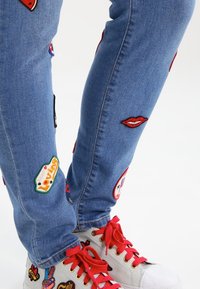 Ljust blå denimjeans med olika färgglada broderade lappar, inklusive läppar och lekfull text, i kombination med grå sneakers med röda snören.