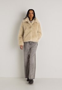 Fuskepelsbeige jakke med bred krage, paret med lysegrå vide bukser. Strukturert stoff og løs passform gjennom hele. Sorte sko.