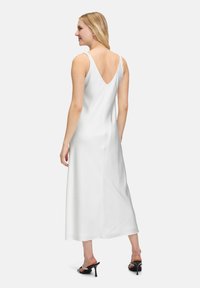 Robe maxi blanche en soie avec un dos en V et des bretelles larges. Présente une texture lisse et tombe jusqu'aux chevilles, accompagnée de talons ouverts noirs.