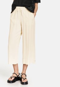 Pantalons large beige clair avec une taille élastique et un cordon de serrage. Le matériau semble lisse et fluide, se terminant juste au-dessus des chevilles.
