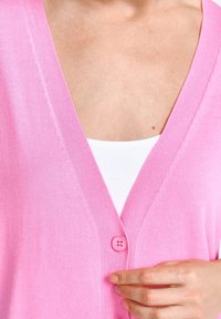 Cardigan rose avec un décolleté en V, texture en maille douce et fermeture à boutons. Assorti à un sous-pull blanc en dessous.