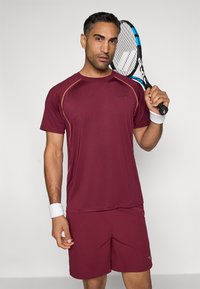 Κάτι χρώματος βουργουνδί αθλητικό μπλουζάκι με αντίθετες ραφές και σορτσάκι, κρατώντας μία ρακέτα τένις. Υφασμάτινη επιφάνεια, σχεδιασμένη για αθλητική απόδοση.