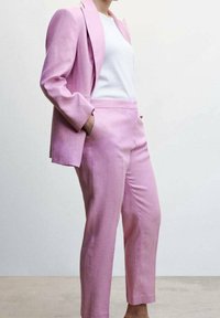 Frau in einem hellroten Anzug mit Blazer und maßgeschneiderten Hosen, kombiniert mit einem schlichten weißen Hemd, steht mit den Händen in den Taschen.