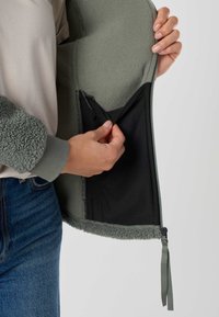 Hände halten eine offene grüne Fleecejacke, die die schwarze Innentasche zeigt, getragen über einem beigen Top und blauen Jeans.