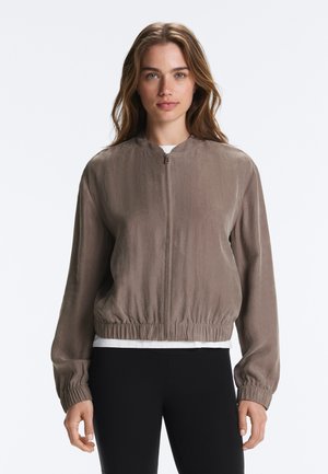 Young woman with loose wavy hair wears a taupe zip-up jacket with elastic cuffs and hem over a white top and black pants.