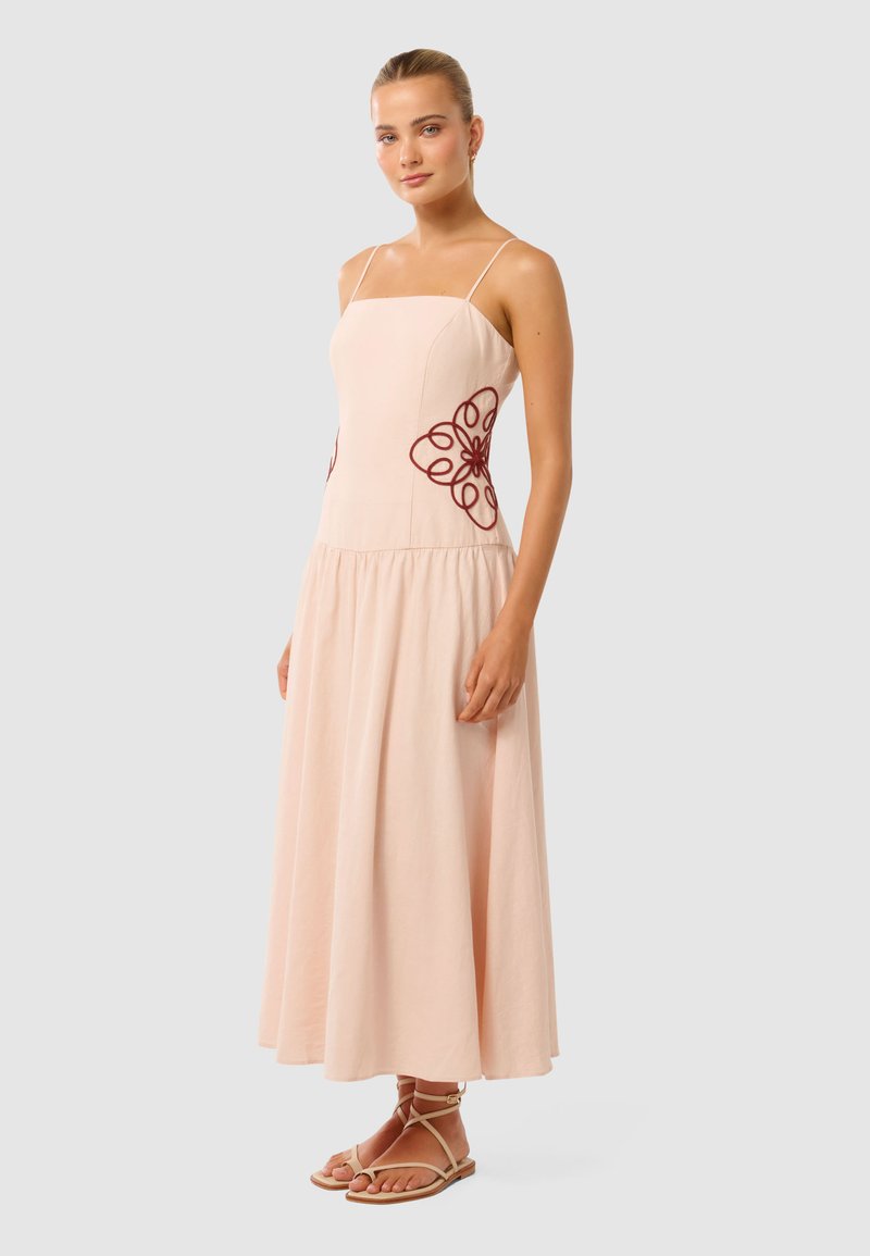 Lichtroze A-lijn jurk met spaghettibandjes en geborduurde bloemenaccenten in kastanjebruin aan de zijkanten, gemaakt van een lichte stof.