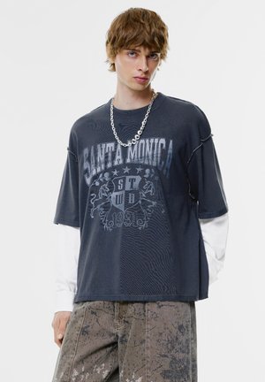 Dunkelgraues, übergroßes T-Shirt mit „SANTA MONICA“-Grafik, kurzen Ärmeln und distressed Details, über einem weißen Langarmshirt getragen.