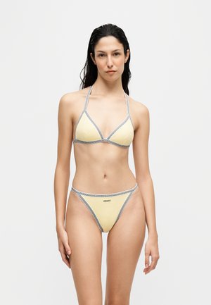 Femme aux cheveux foncés et mouillés portant un bikini jaune clair avec des bordures grises et le logo Versace sur le bas, debout devant un fond blanc uni.