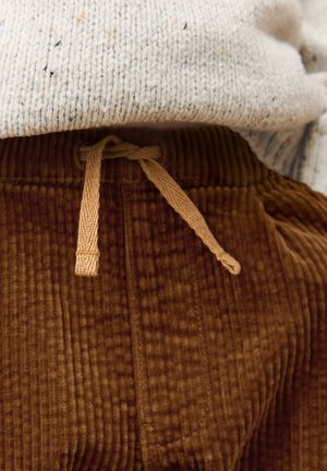 Pantalones de pana marrón con costillas verticales, con cintura ajustable de cordón a juego y una textura suave. Tejido de punto gris parcialmente visible en la parte superior.