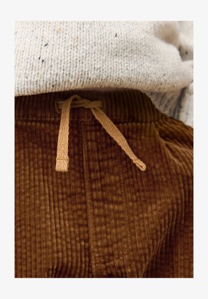 Pantalon en velours côtelé marron avec nervures verticales, doté d'une taille élastiquée avec cordon de serrage assorti et d'une texture douce. Tissu tricoté gris partiellement visible au-dessus.