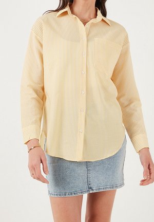 Vrouw draagt een overhemd met lange mouwen, lichtgele strepen en een borstzak, gecombineerd met een vervaagde blauwe denim minirok.