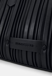 Borsa a tracolla in pelle nera con pieghe verticali, dotata di una toppa con logo impresso e dettagli hardware rotondi. Texture liscia con design strutturato.